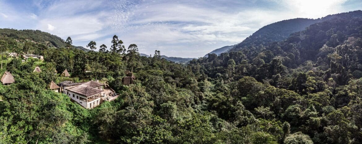 A panoramic view of Bwindi Impenetrable Forest with rolling green hills and a small village of thatched-roof houses nestled among trees under a blue, cloud-dotted sky.