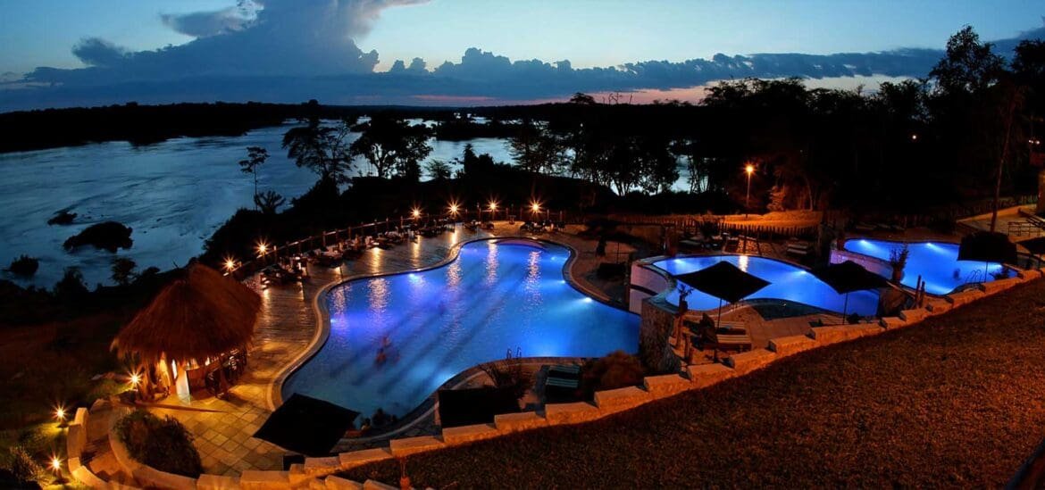 A luxury lakeside resort at night, featuring glowing blue swimming pools, illuminated walkways, and palm trees reflecting in the water of the Nile River.