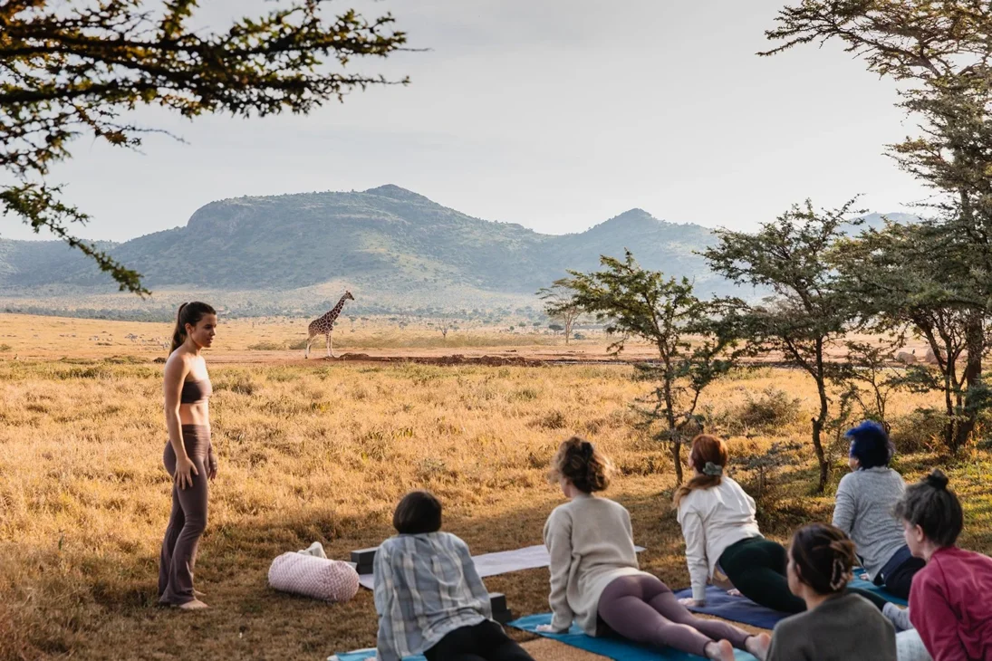 Group practicing yoga outdoors in the African savannah with a giraffe in the distance, blending safari and wellness retreat experience.