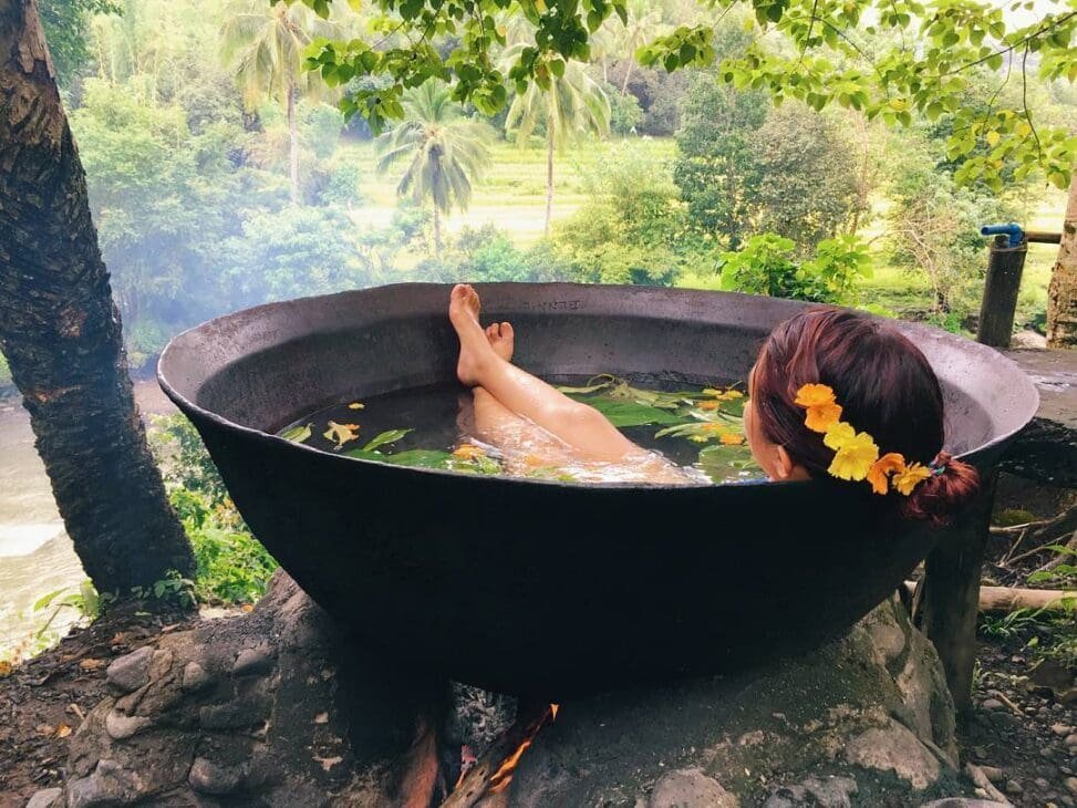 Woman relaxing in a traditional outdoor herbal bath heated over fire, surrounded by lush tropical forest and greenery in the Philippines.
