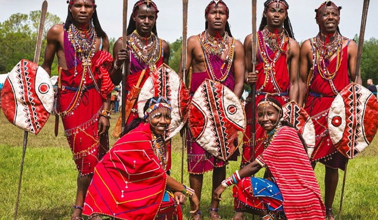 Group of Maasai people in red traditional attire with beaded jewelry, standing and kneeling in a grassy field, holding spears and shields; green trees in the background.