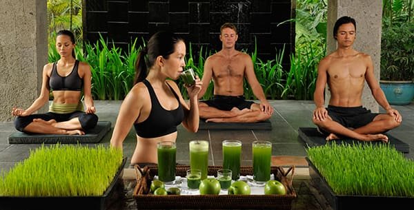 Group of people practicing yoga in a tropical retreat setting with a tray of green juices and fresh produce in the foreground.