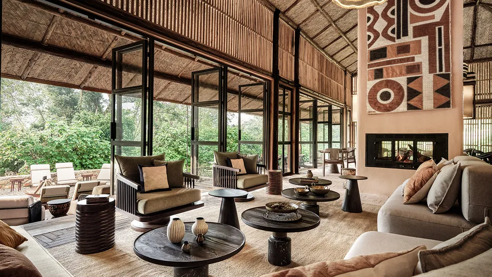 Interior of a luxurious lodge with plush seating, dark wood tables, and a large fireplace; outside, lush greenery is visible through tall black-framed windows, creating a warm, inviting atmosphere.