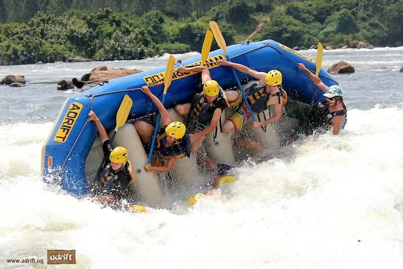 A group of people wearing yellow helmets and life jackets raft a blue and white inflatable boat through white-water rapids on Uganda’s Mighty Nile; lush green banks in the background.
