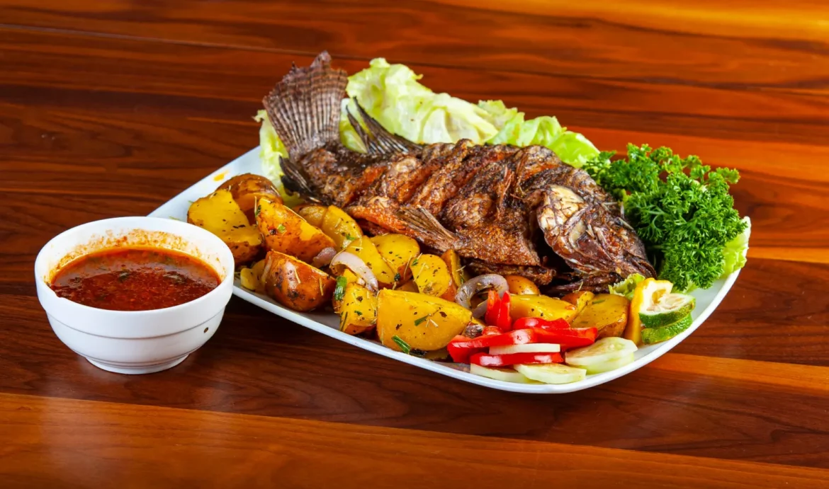 Grilled tilapia served with roasted potato marbles, fresh vegetables, and a small cup of sauce on a plate