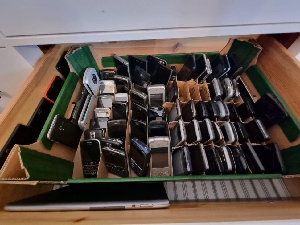 Drawer filled with a large collection of vintage mobile phones neatly arranged on green felt and cardboard dividers, with a silver tablet at the bottom.