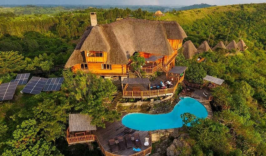 An aerial view of the luxury wood-and-thatch Kyaninga Lodge featuring a turquoise swimming pool and solar panels, perched on a lush green ridge overlooking a crater lake.