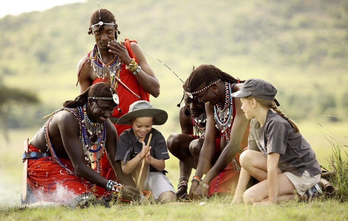 A group of Maasai men in traditional red attire and beaded jewelry gather on a grassy field with two visiting children as they demonstrate starting a fire with sticks; a warm, sunny day in a rural landscape.