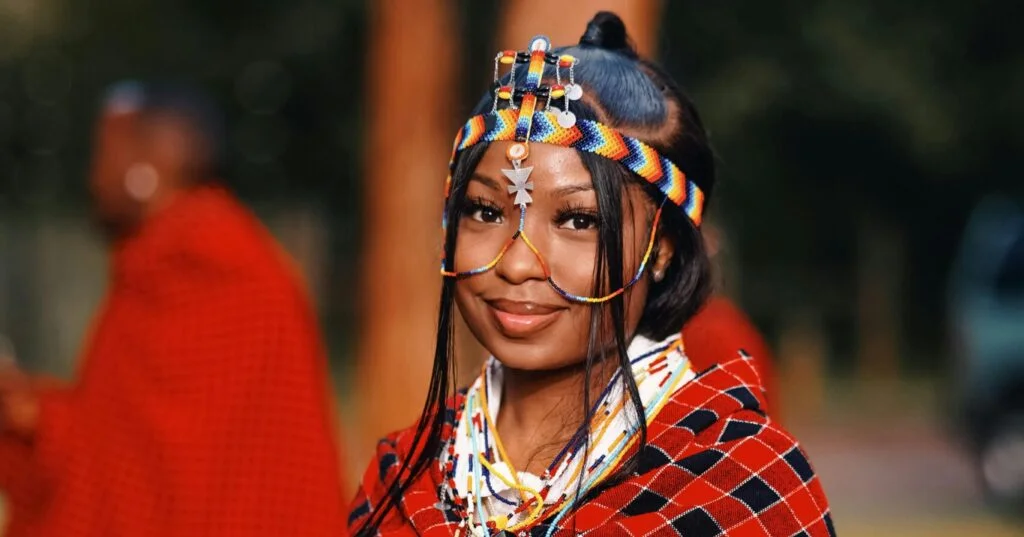 Close-up of a Maasai woman wearing a colorful beaded headdress and necklace, with a slight smile; blurred red-clad figures in the background.