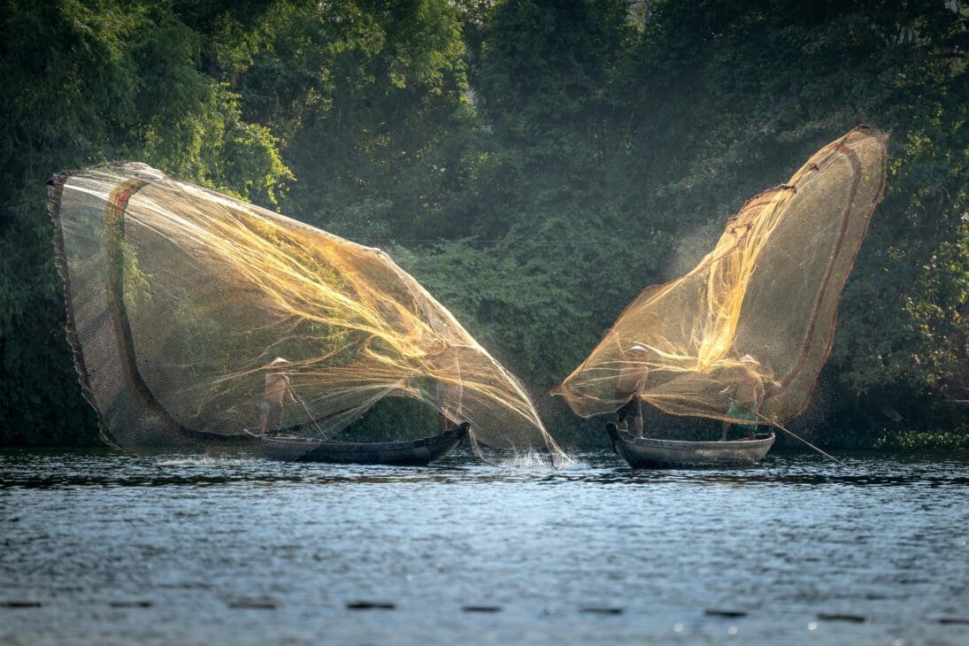 Local fishermen casting nets on Lake Victoria at sunrise, with tourists observing from a boat and birds flying overhead.