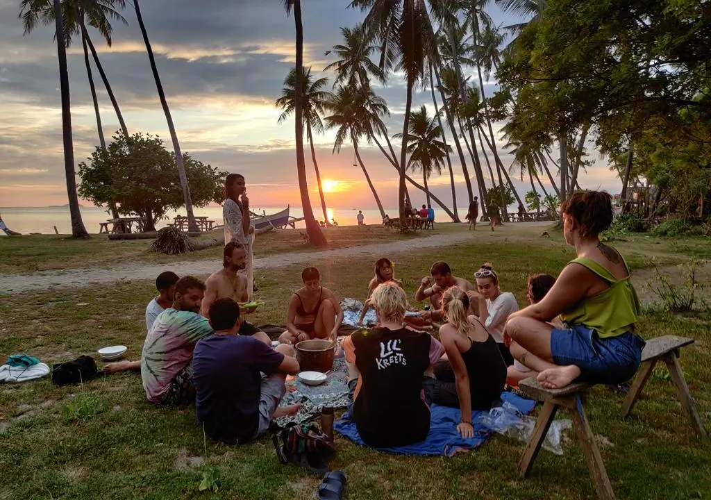 A group of diverse travelers sitting in a circle on a grassy field at sunset, sharing a communal meal on blankets under tall palm trees with a calm ocean view in the background.