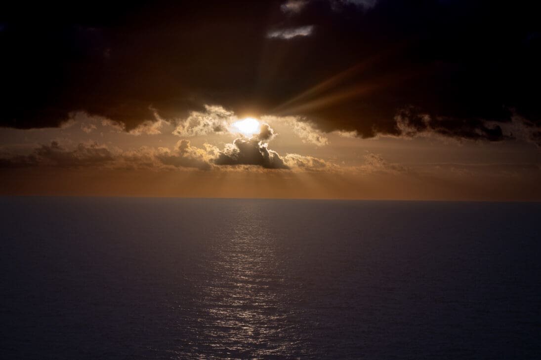 Dramatic sunset over the ocean during a ferry journey across the Philippines, with golden sunlight breaking through dark storm clouds and reflecting across calm sea water.